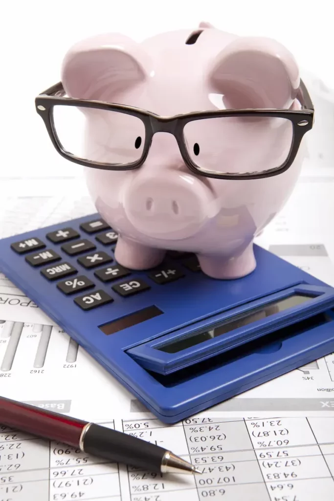 Ein Sparschwein aus Porzellan mit Lesebrille sitzt auf einem Taschenrechner